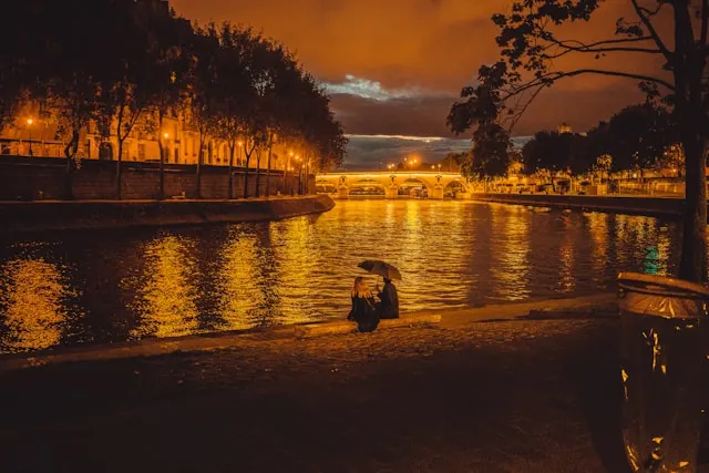 Couple sitting by the Seine River under an umbrella during a rainy night in Paris, near Pont Neuf