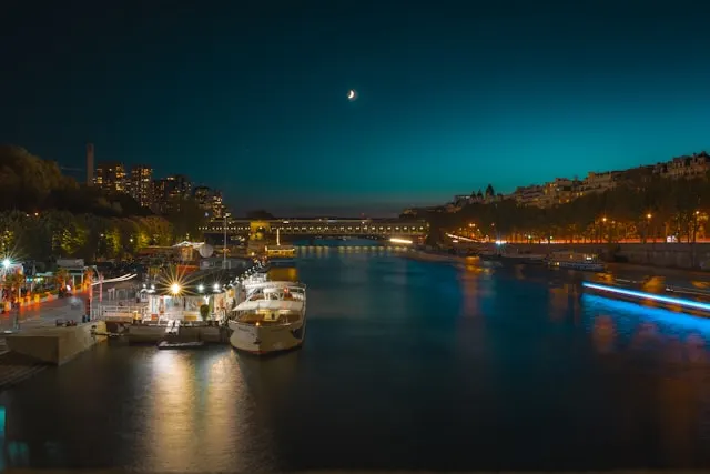 Night view of the Seine River with cruise boats, city lights, and a crescent moon above Paris
